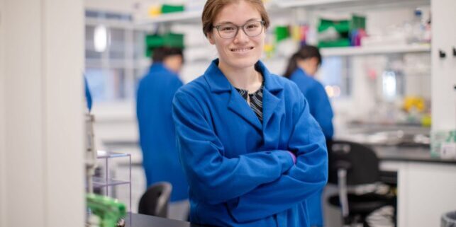 Maikawa in a blue labcoat stands with arms crossed and smiles at the camera. two other researchers are working in the background in the lab.