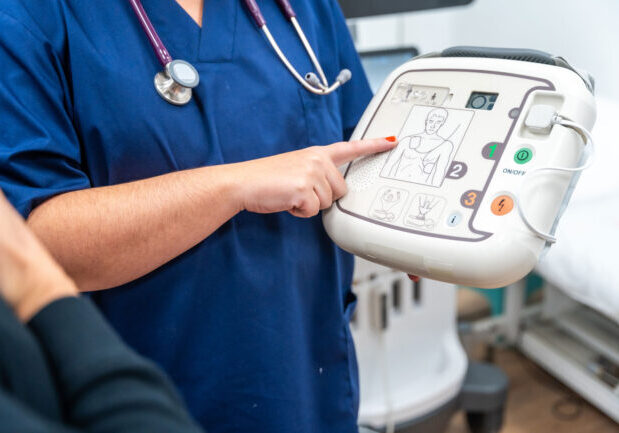 a-cardiologist-explaining-how-a-small-defibrillato-2024-12-09-04-37-01-utc_1500x1000 a medical practitioner wearing a stethoscope points at an AED